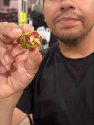 A medium shot shows a man holding a piece of candy in his hand. The candy is cylindrical, with a green exterior and visible red and white fillings. The man's face is partially visible, with a focused expression and a bit of facial hair. He is wearing a black shirt, and the background is blurred, suggesting an indoor setting.