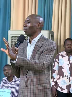 A medium shot captures a Black man speaking into a microphone, gesturing with his hands. He's wearing a checkered blazer over a lighter-colored shirt. Two other men are partially visible in the background. One on the right is wearing a patterned shirt; the other, on the left, is wearing a checkered shirt. A large, white, and gray appliance is behind the main speaker with light blue curtains on either side of it.
