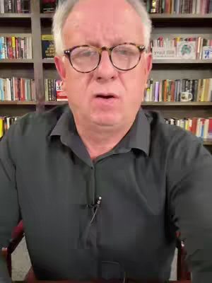 A middle-aged man with glasses is centered in front of a full bookshelf. He wears a dark, button-down shirt and looks directly at the camera with a serious expression. The background is filled with neatly arranged books.