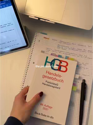 The image shows a well-lit, top-down view of a workspace. A partially opened laptop and a tablet are in the background. In the foreground, there's a hand holding a book titled 'Handelsgesetzbuch' and a pen is lying next to a notebook with visible notes. The overall composition suggests a study or work environment.