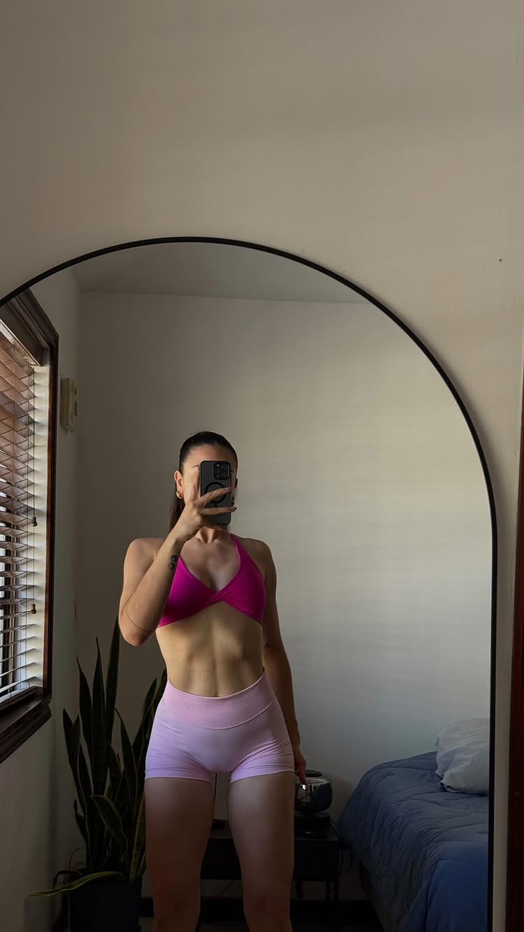 A mirror selfie showing a woman's reflection. She is wearing a pink halter top and light pink shorts, highlighting her well-defined abs. She's holding a phone up to her face, obscuring her features, with a tattoo visible on her arm. The background includes a window with blinds, a houseplant, a bed, and a large, arched mirror.
