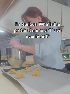 A woman is in a kitchen, behind a counter, in the midst of baking. The scene is brightly lit with neutral-toned walls and cabinets. She appears to be mixing something in a metal bowl and placing dough portions on a baking sheet with a tray of cookies already prepared.