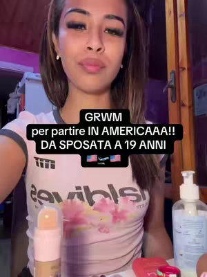 A young woman is filming herself, holding the camera in front of her, with a makeup counter and room in the background. She is wearing a pink t-shirt with a graphic, and there is text overlay. Cosmetics are places in front of her.