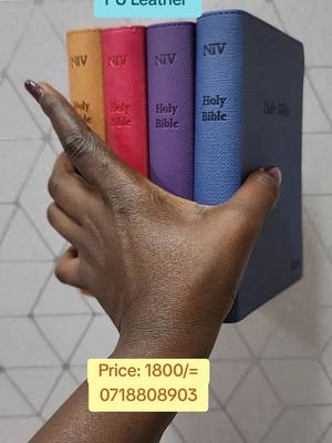 A hand holding a stack of four Holy Bibles in different colors: orange, pink, purple, and blue. The Bibles have white text on their spines; the hand is dark-skinned and holding the books against a light-colored wall. There is also a yellow box with white text which contains a price and a phone number.