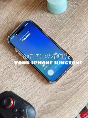 A wooden desk with an iPhone on it is the main focal point. The iPhone’s screen displays a blue background with the words “WhatsApp Audio… Tim Cook”. The screen also shows a call interface, suggesting an incoming call. Overlain on the image is the text “HOW TO CUSTOMIZE YOUR IPHONE RINGTONE”. A black game controller is positioned on the left side of the frame, and a small light blue item sits on the right corner. The overall composition is well-lit, giving a clean and modern feel.