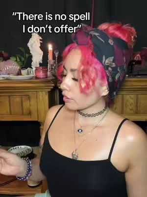 A woman with pink hair and a headscarf is in front of a decorated mantelpiece. The text overlay reads, "There is no spell I don't offer."