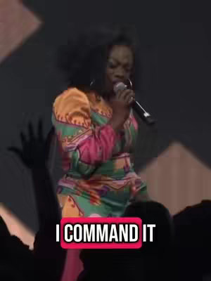 A woman in a colorful dress holds a microphone and sings on stage. A text overlay reads "I COMMAND IT". In the background, the upper bodies of people, possibly an audience, are visible. The lighting is dim.