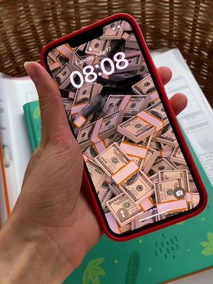 A hand holding a red iPhone. The phone screen displays a wallpaper of stacks of hundred-dollar bills. The background is a mix of a woven surface and pages, suggesting a desk setup.
