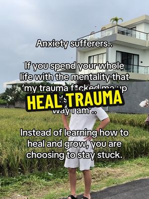 The image shows a man standing in front of a large house with a field in the foreground and a cloudy sky. The foreground has grass and some bushes. The man is wearing a white t-shirt, white shorts, and black sandals. Overlaid on the image is text relating to healing from trauma and advice for anxiety sufferers in a white font with black outlines and a yellow background.