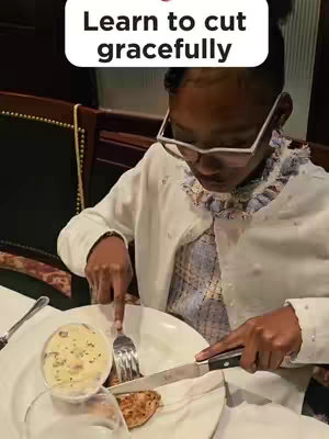 A young person wearing glasses and a white sweater is seated at a table in a restaurant and is in the process of cutting a piece of food on their plate with a knife and fork. The scene is well-lit, and other dishes, silverware, and a partially visible glass are present on the table.