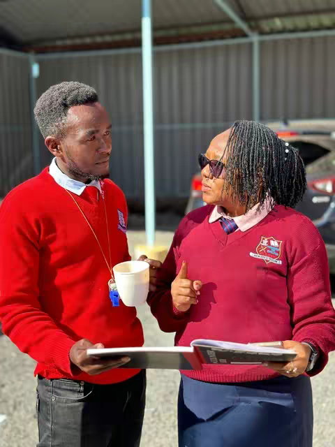 Two people, a man and a woman are standing outside. The man is holding a coffee cup and a pad of paper, with a whistle hanging from his neck. The woman is pointing. They are wearing school uniforms.