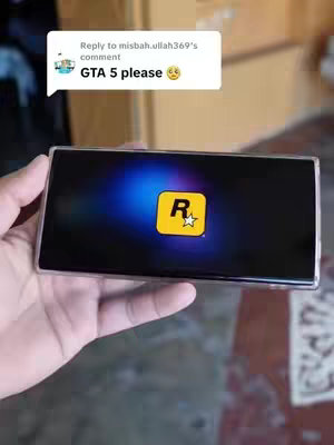 A hand holding a phone displaying the Rockstar Games logo. The background is blurred, suggesting an indoor setting.