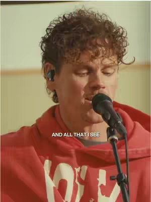 A medium shot shows a light-skinned man singing into a microphone. He wears a red hooded sweatshirt and has curly brown hair. Text overlayed on the image reads, 'AND ALL THAT I SEE.' The background is a blurred light color.