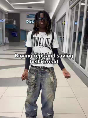 A young man with dreadlocks and sunglasses stands in what appears to be a mall, wearing a white t-shirt with a black logo, distressed jeans, and a backpack. The background shows glass windows and retail storefronts.