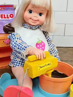A close-up shot of a vintage 1973 Hasbro Sweet Cookie doll. The doll has blonde hair, blue eyes, and freckles, wearing a bib with a strawberry image, and a blue checkered dress. She is holding and using a yellow toy mixer, positioned next to a bowl with a dark brown substance. A purple box of Jell-O chocolate pudding is partly visible in the background, next to a small flower-shaped decoration.