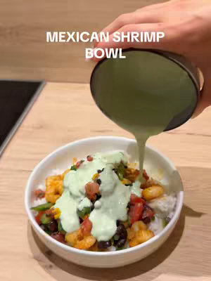 A close-up shot of a white bowl filled with ingredients for a mexican shrimp bowl. A hand is pouring a green creamy sauce over the bowl. The background is a wooden table.