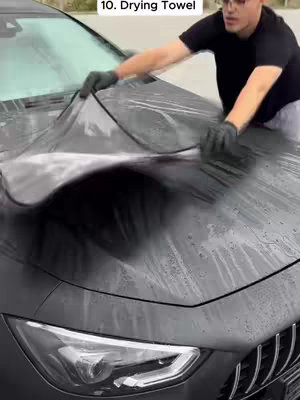 A man is wiping a black car with a large drying towel. The towel is draped over the car's hood, and the man is holding either end of the towel. The car's hood is wet, and there is a close-up of the front of the car, emphasizing its features and design.