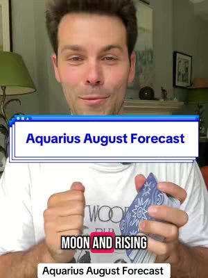 A man with short brown hair is centered in the frame, smiling and looking at the camera. He is holding a deck of cards in one hand. Above his head, a blue rectangle with white text reads 'Aquarius August Forecast'. Below him, another text box with the words 'Moon and Rising' is shown. The background includes a green wall, a light and other home decor.