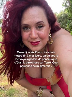 A selfie-style image featuring a woman with long, red hair and bold red lipstick. She wears a low-cut top. A text overlay appears beneath her, partially obscuring her chest. The background features lush greenery, suggesting an outdoor setting. The overall composition is slightly overexposed.