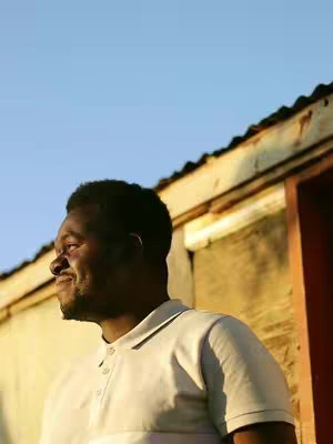 A man in a light-colored polo shirt stands in front of a rustic building with a blue sky in the background. He is looking towards the sky with an expression of joy on his face.