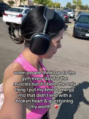 A woman with dark hair pulled back in a bun is shown from the side, wearing headphones and a pink workout top, walking past parked cars. Text is overlaid on the image.