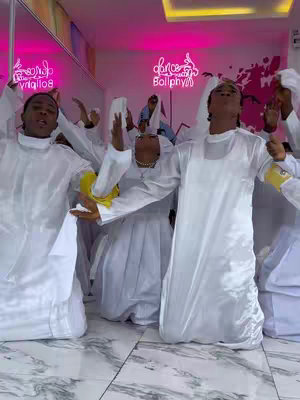 A group of people dressed in white robes with raised hands, dancing or performing a routine. The setting appears to be a music studio with a pink neon sign.