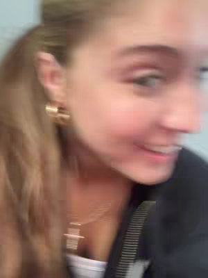 A slightly blurred selfie featuring a young woman with light hair pulled back in a ponytail. She is wearing gold hoop earrings and a delicate gold necklace with a cross pendant. She is smiling with a joyful look. The background is out of focus.