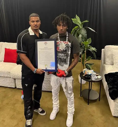 The image captures two men posing indoors. On the right is a man with dark dreadlocks, wearing a dark green t-shirt with patterned design, white distressed jeans, and white sneakers, with large jewelry. He is holding a framed document with a blue border.  To his left stands a man in a black shirt, black pants, and black and white sneakers, holding the other side of the frame. The background includes a white couch, a plant, and a black curtain.