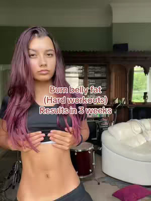 A woman with purple hair stands in front of a background, displaying her abs and holding her stomach in front of the camera. Text overlayed on the image states how to get rid of belly fat in 3 weeks with "hard workouts."