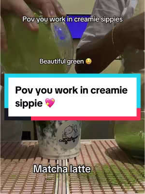 The image shows a person working in a cafe setting. The shot is taken from a first-person perspective, with hands holding a glass of bright green liquid. The image has multiple text overlays.