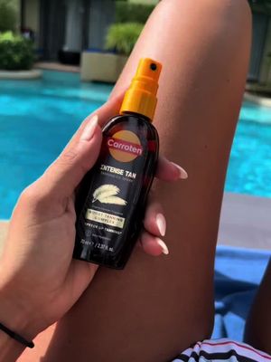 A woman's arm and hand are shown holding a small bottle of Carroten tanning oil. The background is a blurred view of a swimming pool. The bottle is black with the Carroten logo and product information visible.