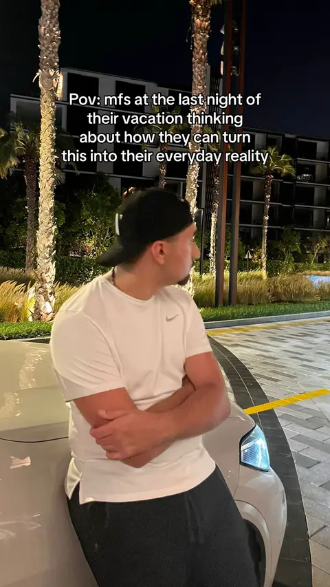 A night shot featuring a man leaning against a silver car. The man is wearing a black cap, a white shirt, and dark pants. The background includes high palm trees and modern buildings under a dark sky, suggesting an evening out in a resort or urban setting.