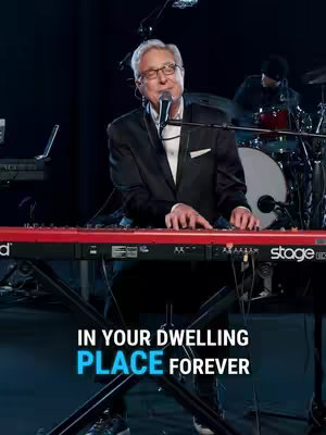 A man playing a red keyboard on a stage, with a microphone in front of him and words overlaid on the bottom of the image. In the background, there's a drummer and other stage equipment.