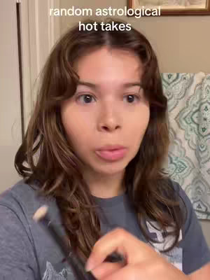 A woman with brown hair and light skin is centered in the frame, holding a makeup brush. She's giving a slightly surprised or thoughtful expression while looking at the camera. In the background, a patterned shower curtain and a glimpse of a framed picture are visible.