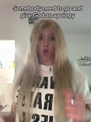 A woman with long blonde hair has her mouth open in shock or surprise. She is wearing a white shirt with a graphic on it.