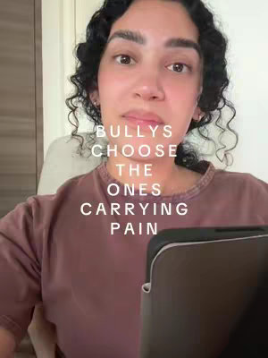 A medium shot of a woman with curly black hair looking directly at the camera. She appears to be inside, likely in an office, with a neutral background. Overlaid on her face is text that reads 'BULLYS CHOOSE THE ONES CARRYING PAIN'.