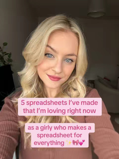 A young woman with long blonde hair and blue eyes is smiling at the camera. She's wearing a brown ribbed shirt and a light lilac color block shapes text on her chest. The text reads "5 spreadsheets I've made that I'm loving right now" and "as a girly who makes a spreadsheet for everything ✨🎀💖". The background is a blurred interior with hints of a window and white furniture.
