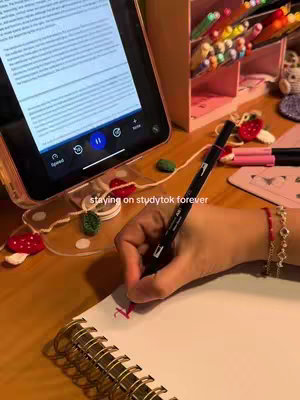 The cover image features a close-up view of a workspace, likely a desk. A tablet is propped up, displaying text, with a hand holding a pen, writing in a notebook.  There is a pen holder with pens of various colors on the right, with other desk items.