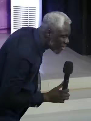 A medium shot of a man with white hair and a dark shirt holding a microphone and speaking with emotion. He is in what appears to be a stage or auditorium setting.