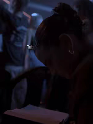 A person, viewed from the back and slightly to the side, is reading from a document with a blurry background behind them. The person, presumably a female, is in focus with her hair styled in a bun. The text 'Tuta' appears as an overlay.