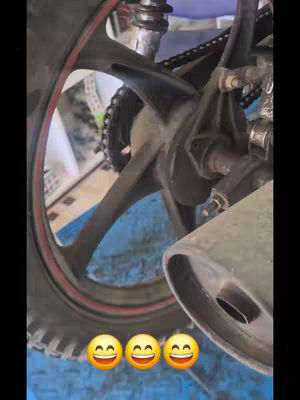 Close-up shot of a motorcycle tire and wheel. The tire has a red stripe, and the wheel has black spokes. The background is blurred, but appears to be a blue surface. Three smiling face emojis are at the bottom