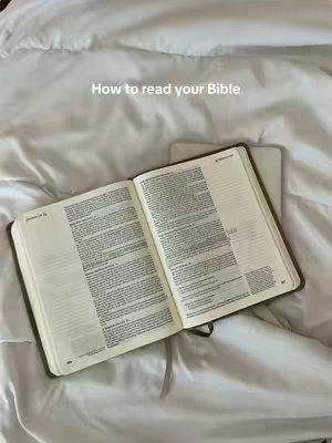 An open book lying on a bed with white sheets. The book is in the center, with its pages showing text. Above the book's center, the text 'How to read your Bible' is written. The background features the white sheets.