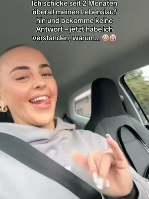 A selfie taken inside a car with a woman speaking directly to the camera. The woman has fair skin, light-colored hair, and is wearing a grey hoodie and gold hoop earrings. She's smiling and sticking her tongue out. There is text overlayed on top of the image.