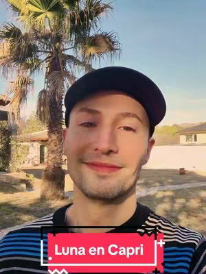 A man wearing a black baseball cap and a striped shirt smiles towards the camera against a blurred background of palm trees and building exteriors. A red and white box in the foreground contains the text 'Luna en Capri'.
