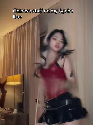 A woman with dark hair and a white front section is dancing in what appears to be an apartment or home setting. She's wearing a red top and a black skirt. The background includes neutral-colored curtains and a lamp.