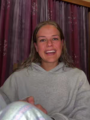 The image shows a young woman smiling directly at the camera, wearing a gray hoodie. Her blonde hair is parted on the side and falls to her shoulders. Behind her are reddish and white patterned curtains. The lighting is soft, and the composition is a medium shot.