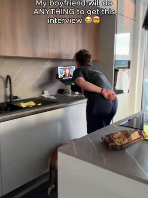A man stands at a kitchen counter, facing a laptop that appears to be displaying a video call. He is turned away from the camera with his back to the viewer. The kitchen has wooden cabinets on top and a modern black counter.  The caption on the image reads,  "My boyfriend will do ANYTHING to get this interview".