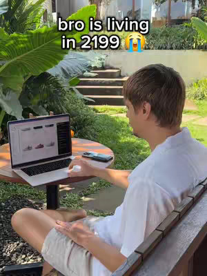 A man is seated outside, working on a laptop. The background features greenery, a water feature, and a small table. The man is seen from the back, wearing a white shirt.