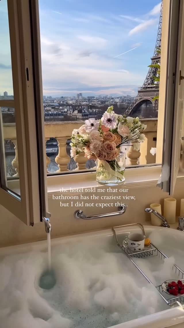 The cover image shows a luxurious bathroom in a hotel suite. The focus is on a large bathtub filled with foamy water, with a partially visible view of the Eiffel Tower and Parisian cityscape framed by an open window. A vase of flowers sits on the windowsill, and a tray with a cup, cookies and berries rests on the side of the tub.
