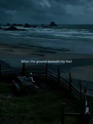 A moody, wide shot of a beach scene under a cloudy sky. The ocean stretches out, meeting a rugged coastline with dark rocks. In the foreground, there's a wooden fence and a small table with a figure seated with their back to the camera, further contributing to the pensive atmosphere and evoking a sense of solitude. The sky is a dominant feature, filled with shades of gray, reflecting a sense of tranquility.
